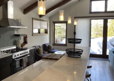 A modern kitchen with a marble island, hanging pendant lights, and wooden beams on the ceiling, with a view of a snowy landscape through the glass door.